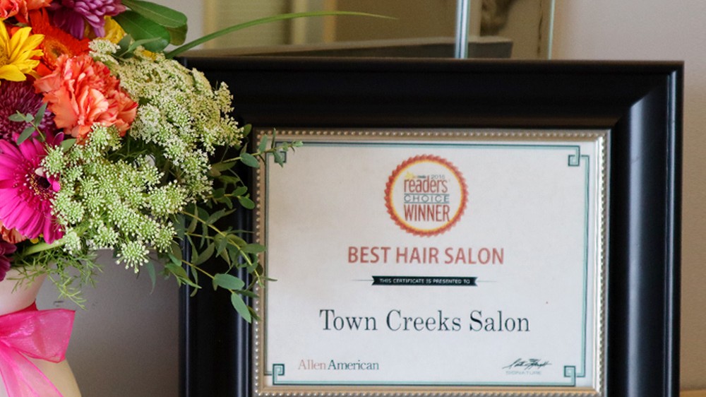 A framed certificate awarded to Town Creeks Salon as the Best Hair Salon by Allen American's Reader's Choice sits proudly on a wooden surface, next to a colorful bouquet of flowers tied with a pink ribbon. The salon, which offers leasing options for stylists, celebrates this community recognition.